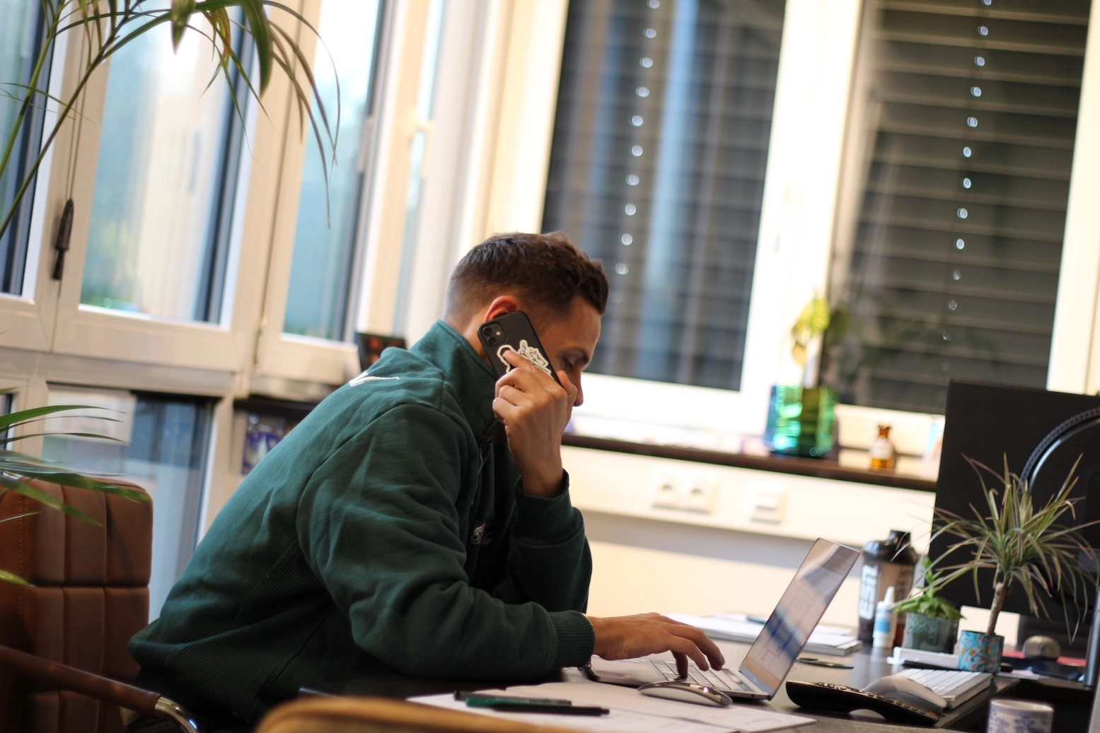 „Geschäftsführer Basti arbeitet konzentriert an seinem Laptop und telefoniert gleichzeitig im Büro.“