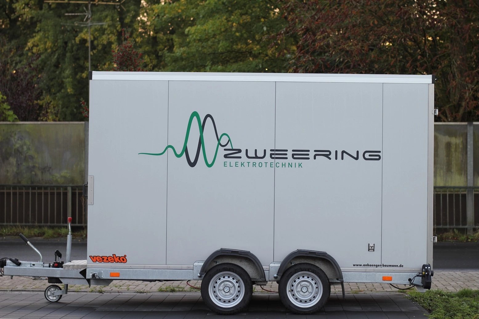„Transportanhänger mit Zweering-Elektrotechnik-Logo auf einem Parkplatz vor einer Grünanlage.“